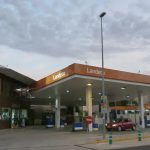 Gas station with orange canopy reading Landete, pumps under a covered forecourt and a red car at a pump under a cloudy sky.