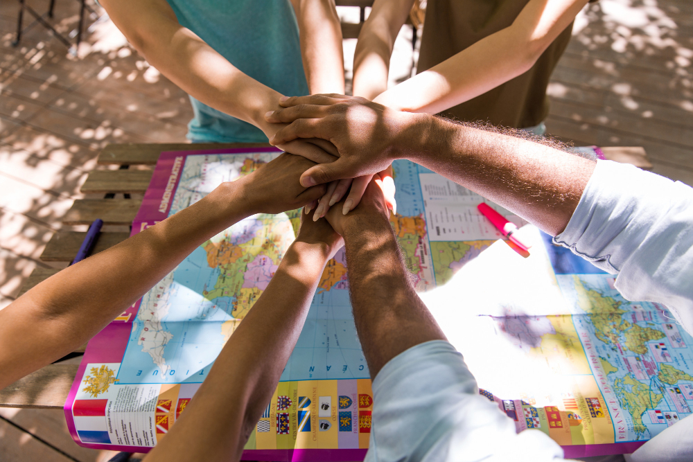 773630 - Fundación Trébol Educación | CR Diario Group of people placing hands together over a world map on a table, symbolizing teamwork in planning travel or a project.