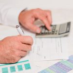 773652 - Carbonell Consultors Associats | CR Diario Person signs a form with a pen while using a calculator, surrounded by financial documents and color charts on a desk
