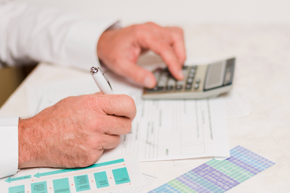 773652 - Carbonell Consultors Associats | CR Diario Person signs a form with a pen while using a calculator, surrounded by financial documents and color charts on a desk