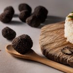 774679 - Cristofaro Trufas | CR Diario Black truffles resting on a wooden spoon beside a round herb-topped rice cake on a rustic wooden board.