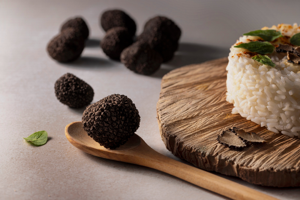 774679 - Cristofaro Trufas | CR Diario Black truffles resting on a wooden spoon beside a round herb-topped rice cake on a rustic wooden board.