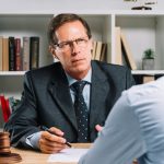 774908 - Ásegas Abogados | CR Diario Lawyer in a dark suit and glasses discusses a document with a client across a wooden desk, a gavel nearby.