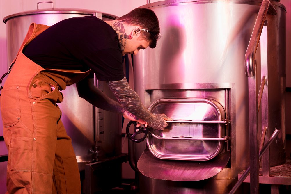 778477 - Multifiltra | CR Diario Worker in orange coveralls and safety glasses opens a large stainless steel industrial kettle door with a hand wheel grip.