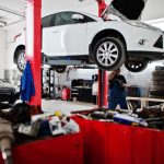 Auto repair shop scene with a white car elevated on a hydraulic lift and two technicians at work in the background.