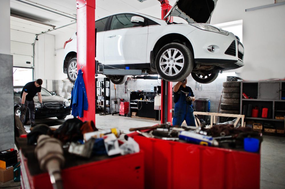 Auto repair shop scene with a white car elevated on a hydraulic lift and two technicians at work in the background.