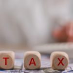 Tax word on wooden cubes with a woman working in the background | CR Diario Wooden blocks spell TAX over a cash-filled surface, with a blurred person using a calculator in the background (tax theme).