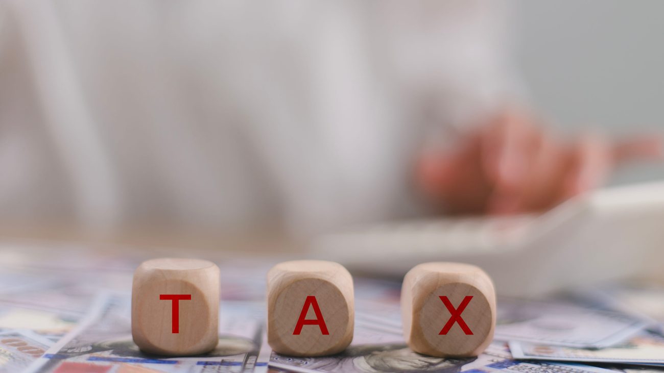 Wooden blocks spell TAX over a cash-filled surface, with a blurred person using a calculator in the background (tax theme).