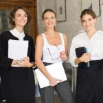 El trabajo de las azafatas fortalece la organización de eventos profesionales exitosos - 779730 - TISA | CR Diario Three professional women posing for a photo in an office, each holding notebooks or a laptop and smiling at the camera in front of a whiteboard.