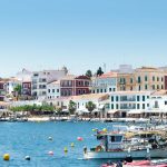 757555 - Menorca Secret | CR Diario Coastal town along a blue harbor with colorful buildings and boats docked by a stone promenade.