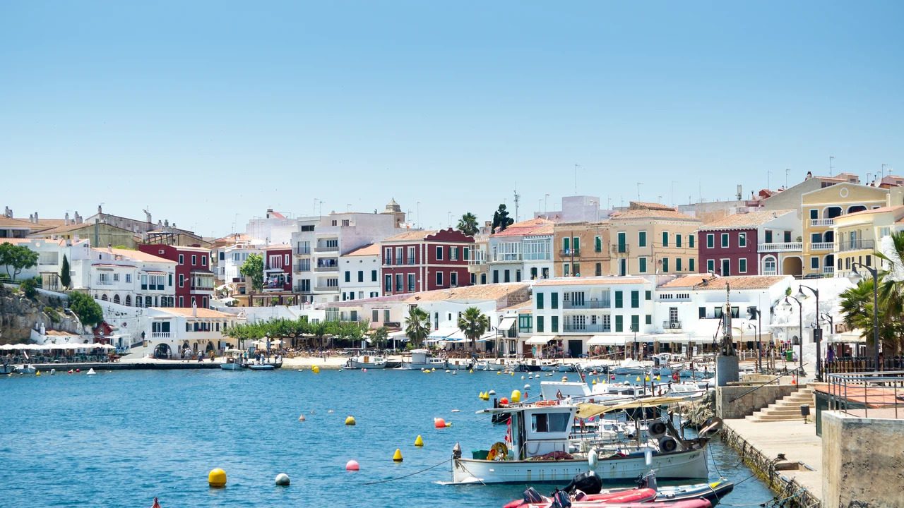 757555 - Menorca Secret | CR Diario Coastal town along a blue harbor with colorful buildings and boats docked by a stone promenade.