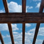 Wooden roof with blue sky and white clouds as a background | CR Diario Wooden beam framework viewed from below against a blue sky with white clouds.
