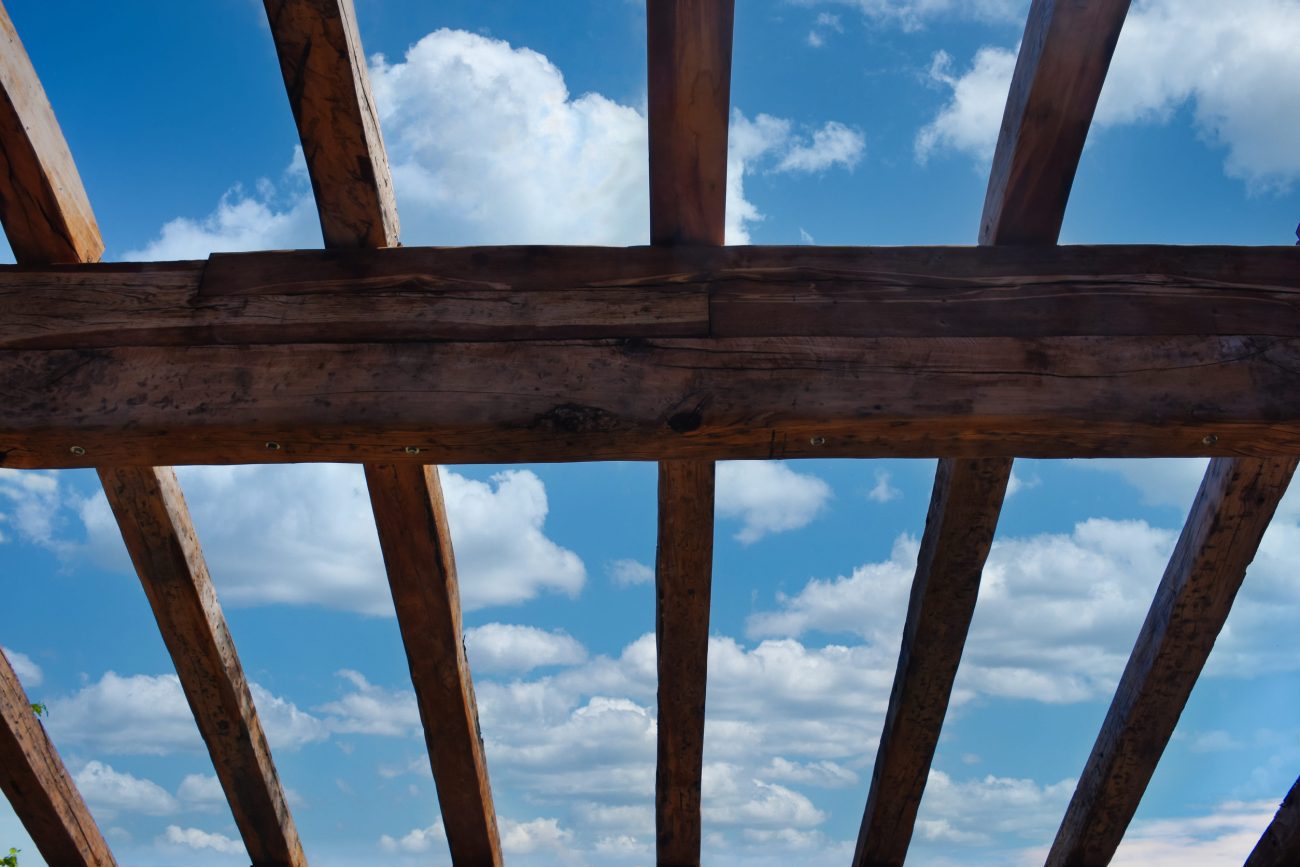 Wooden roof with blue sky and white clouds as a background | CR Diario Wooden beam framework viewed from below against a blue sky with white clouds.