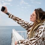 780814 - SeaConnect | CR Diario Person with zebra-print shirt and long dreadlocks extends an arm to take a selfie by the sea, wearing glasses against a blue sky and railing.