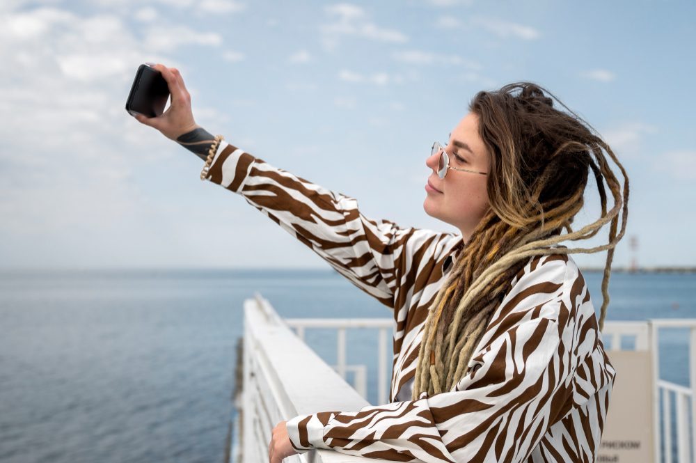 780814 - SeaConnect | CR Diario Person with zebra-print shirt and long dreadlocks extends an arm to take a selfie by the sea, wearing glasses against a blue sky and railing.