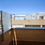 Sunlit urban balcony with glass safety panels and a brick railing, overlooking neighboring rooftops with solar collectors under a blue sky.