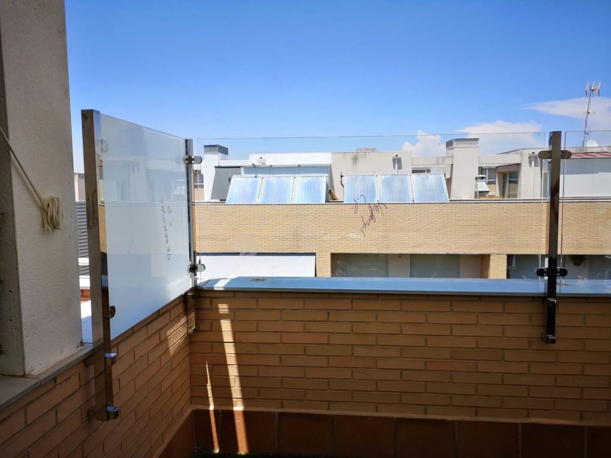 Sunlit urban balcony with glass safety panels and a brick railing, overlooking neighboring rooftops with solar collectors under a blue sky.