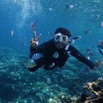 782045 - Brisub | CR Diario Diver in a black wetsuit and mask swims underwater among a colorful reef surrounded by tropical fish, hands forming a peace sign.