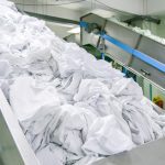 clothes in conveyed to washing machine | CR Diario Industrial laundry facility with a metal conveyor belt piled with white linens