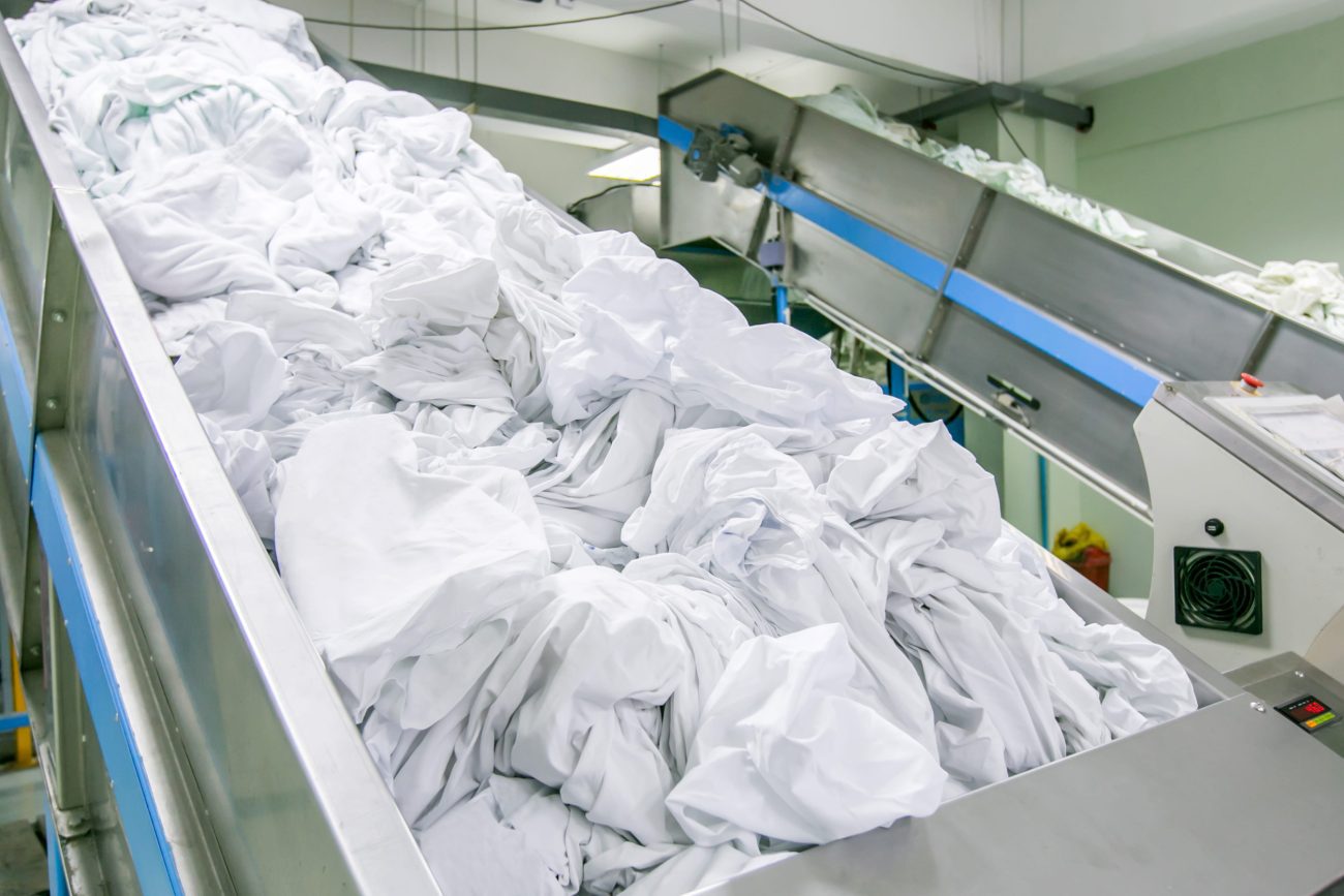 clothes in conveyed to washing machine | CR Diario Industrial laundry facility with a metal conveyor belt piled with white linens
