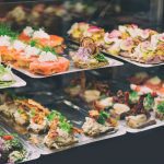 782592 Pagadi Taberna | CR Diario Assorted bite-sized pastries and canapés on metal trays in a display case, featuring smoked salmon, cream cheese, herbs, and colorful toppings.