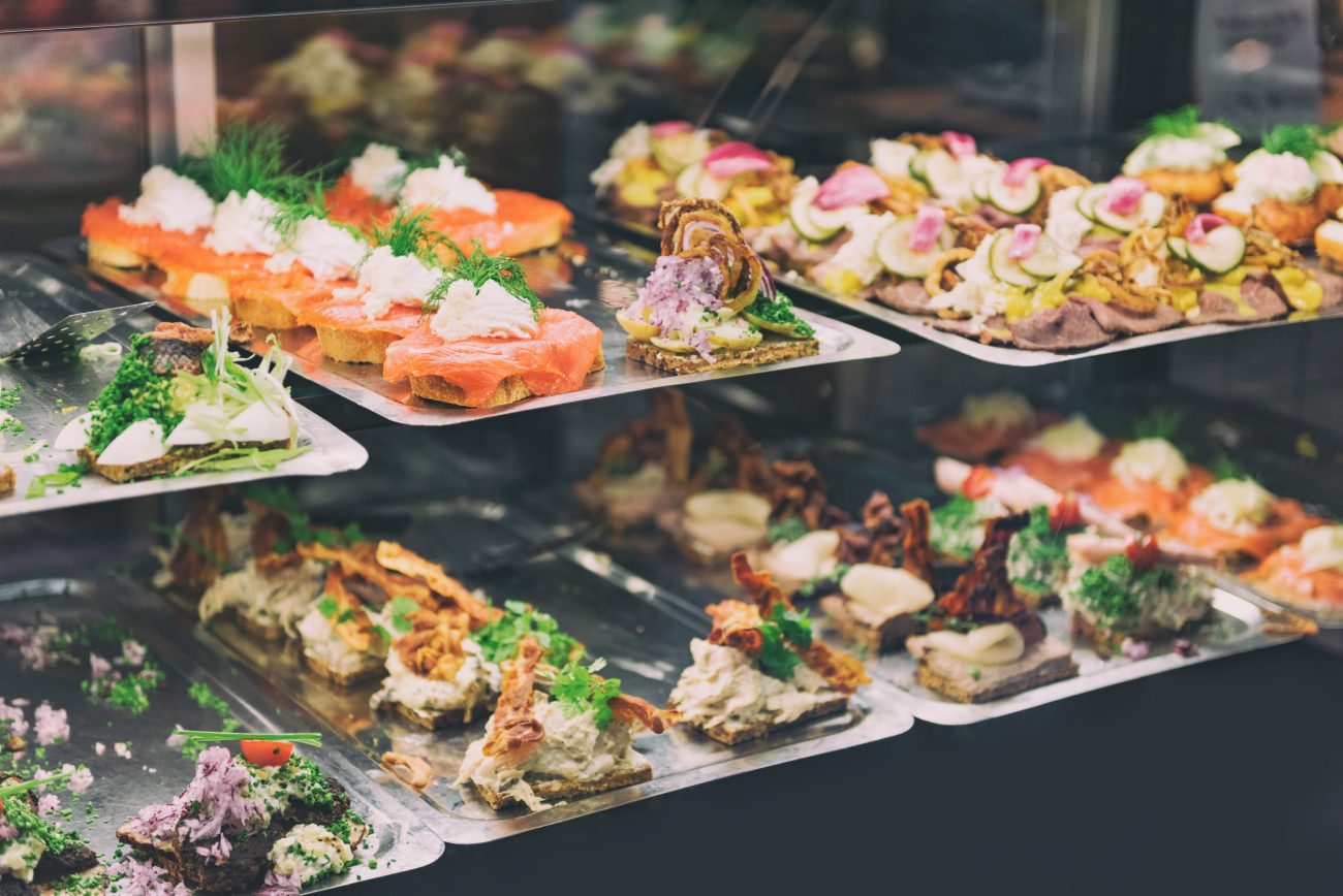 782592 Pagadi Taberna | CR Diario Assorted bite-sized pastries and canapés on metal trays in a display case, featuring smoked salmon, cream cheese, herbs, and colorful toppings.