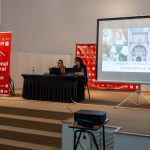 Two presenters sit at a black-draped table on a stage, microphones ready, with a projector screen showing a slide about Torrijos art and a red Bienal banner nearby.