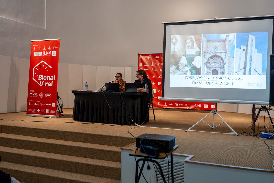 Two presenters sit at a black-draped table on a stage, microphones ready, with a projector screen showing a slide about Torrijos art and a red Bienal banner nearby.