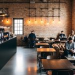 Industrial-style cafe with brick walls and warm pendant lights; barista near the counter while customers work on laptops and read papers.