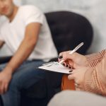 784024 - Sandra Barcenilla Psicóloga | CR Diario Counselor writes on a clipboard during a therapy session, while a man sits blurred in a chair in the background.