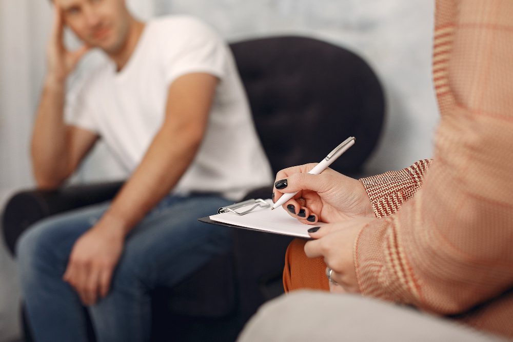 Counselor writes on a clipboard during a therapy session, while a man sits blurred in a chair in the background.