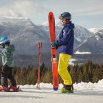 785048 - Erascola | CR Diario Adult in blue jacket and yellow pants holds bright orange skis on a snowy slope, with a child learning to ski nearby.