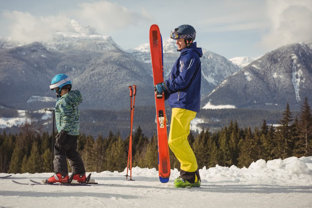 Adult in blue jacket and yellow pants holds bright orange skis on a snowy slope, with a child learning to ski nearby.