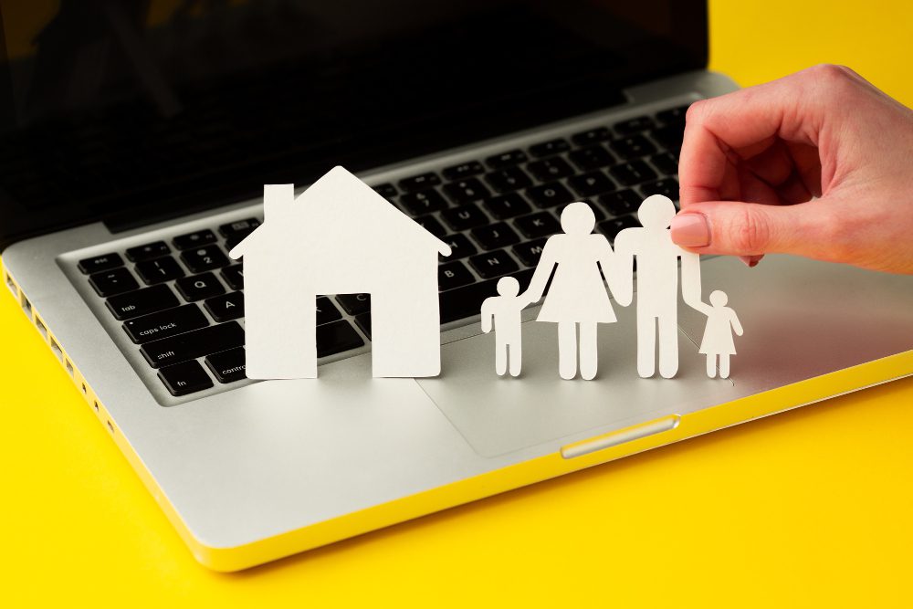 Paper cutouts of a house and a family placed on a laptop keyboard, with a hand arranging the figures, symbolizing online housing or family planning