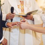 785136 - Cantalapiedra Abogados | CR Diario Bride placing a ring on the groom's finger during their wedding ceremony, with the officiant holding a mic in the background
