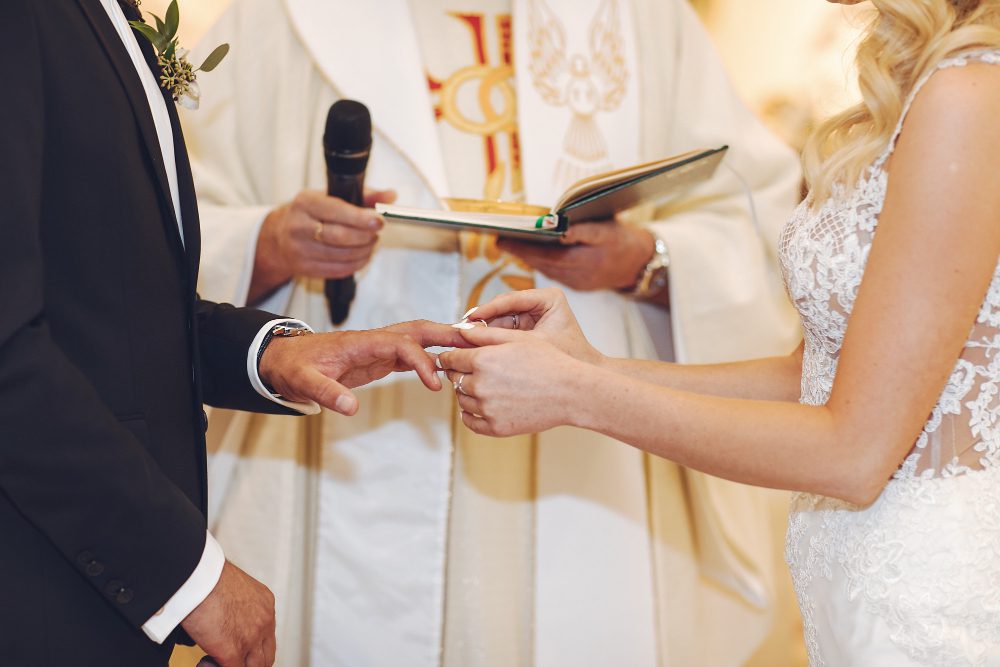 Bride placing a ring on the groom's finger during their wedding ceremony, with the officiant holding a mic in the background